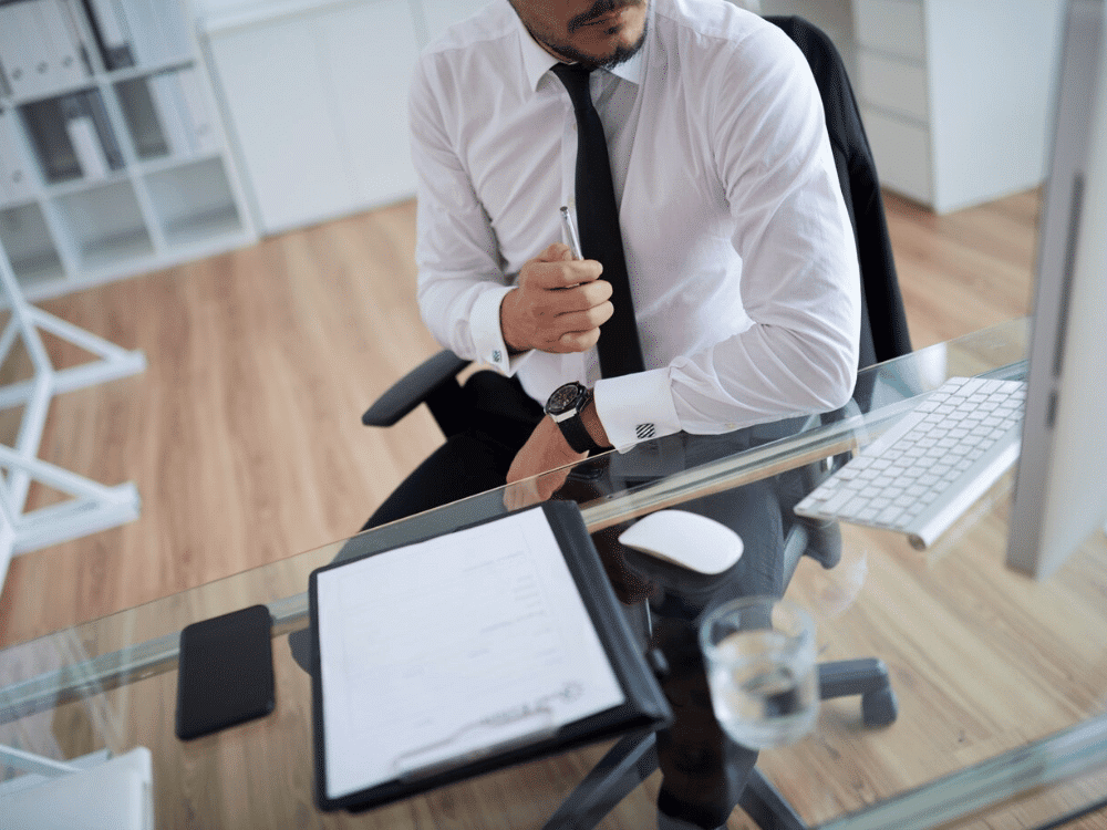 Man in white shirt contemplating at desk with pen and documents.