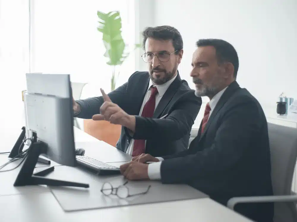 Two businessmen in suits discussing data on a computer screen.